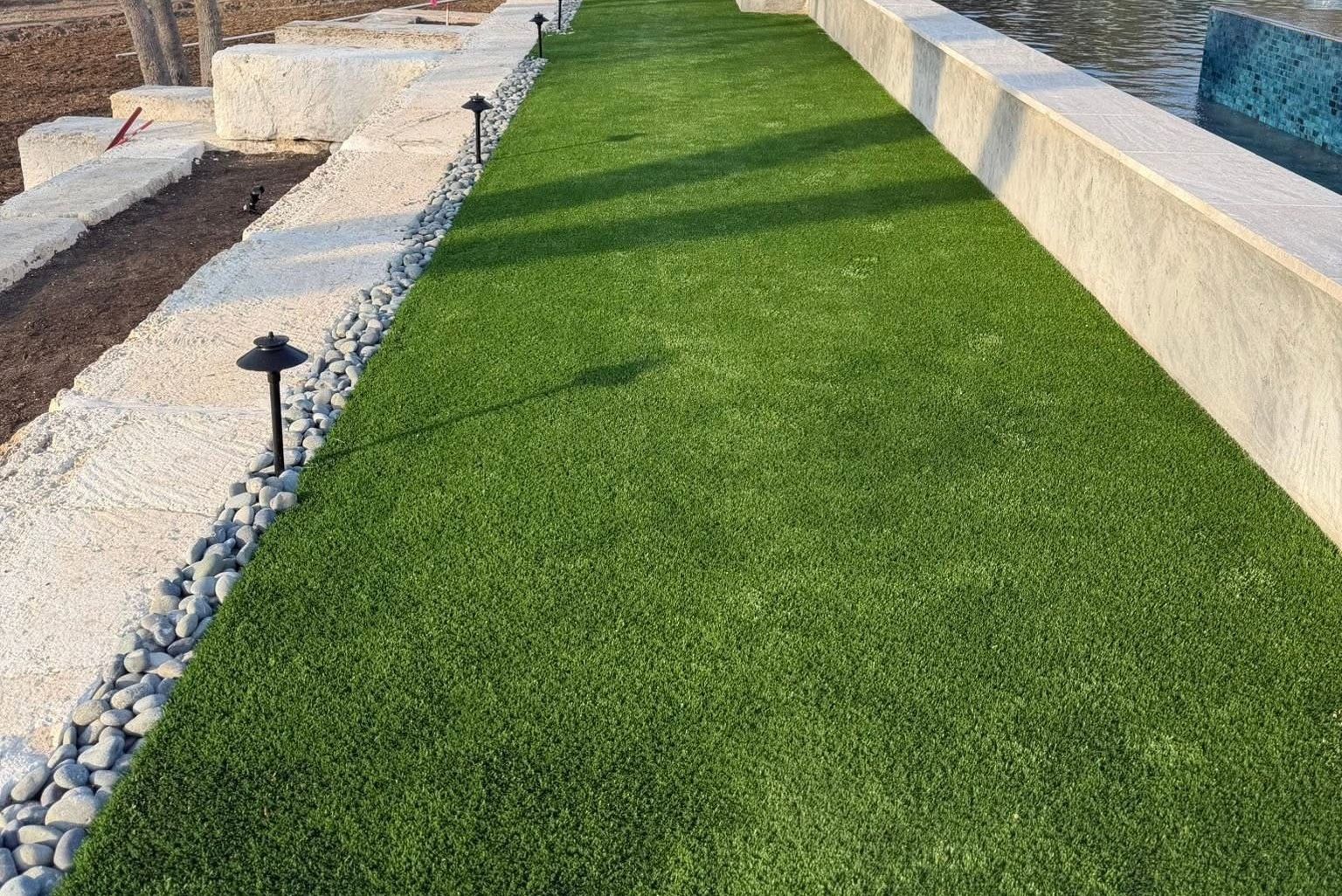 Artificial turf lawn bordered by cut limestone blocks, river rock and path lights beside a raised pool wall